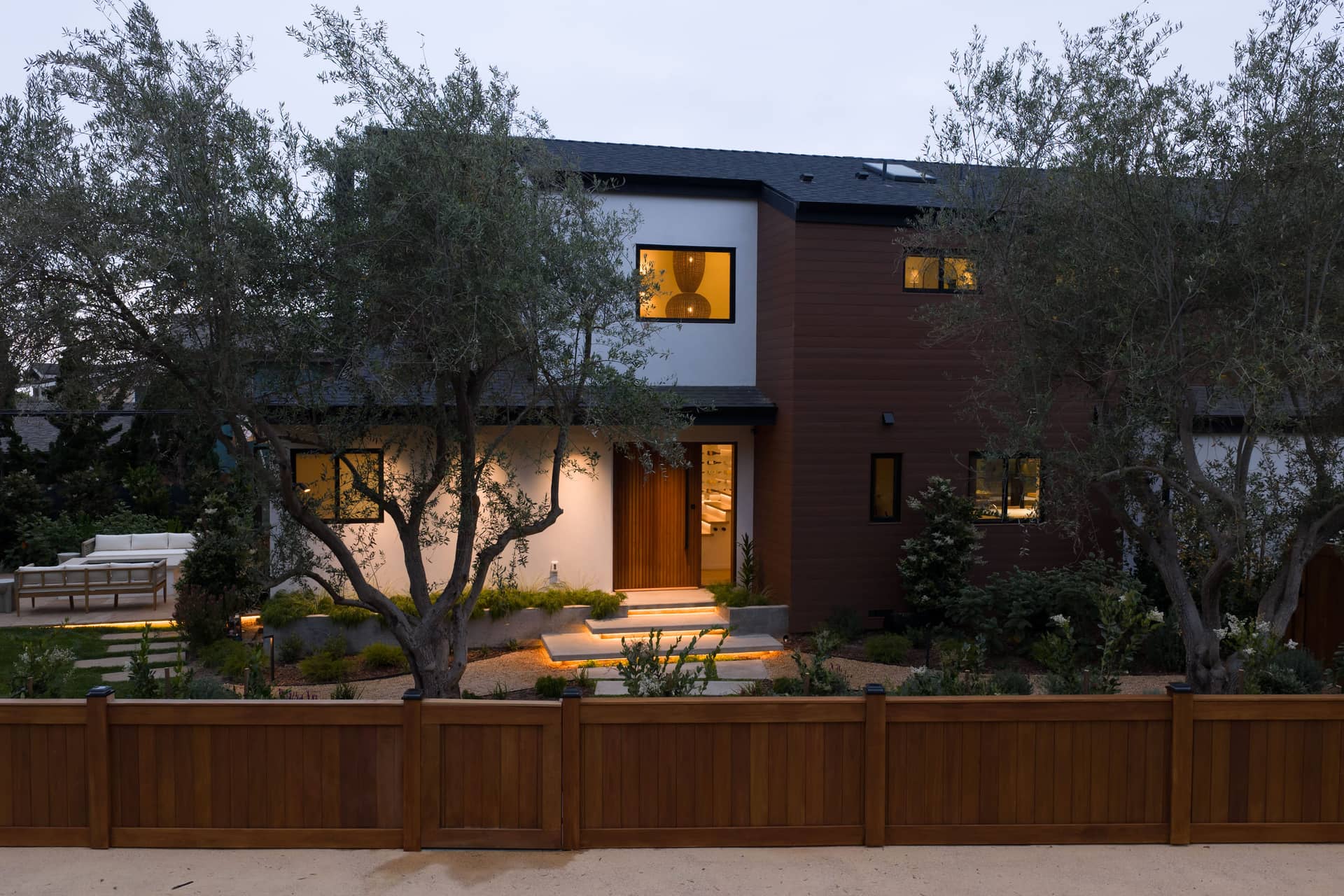 The Padaro Lane residence at dusk, the entry lit behind olive trees and a wood fence.